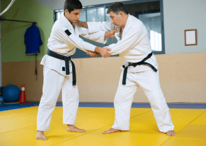 Two judo practitioners in white gis and black belts practicing a throw on a mat in a dojo with a green wall background.