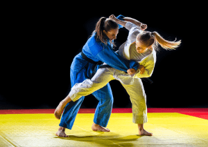 Two female judo practitioners in blue and white gis, performing a dynamic throw on a yellow mat with a black background.