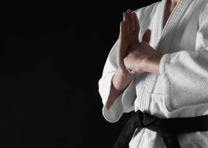 A martial artist in a white gi with a black belt is performing a hand strike isolated against a black background.