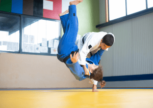 Two judo practitioners in action: one in a white gi performing an over-the-shoulder throw on an opponent in a blue gi.