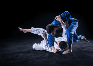 Two judo athletes in action: one in a blue gi executing a ground technique on an opponent in a white gi on a black mat.