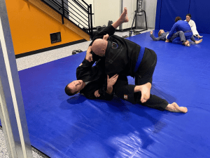 Adult Brazilian Jiu-Jitsu training session with two participants practicing grappling techniques on a blue mat.