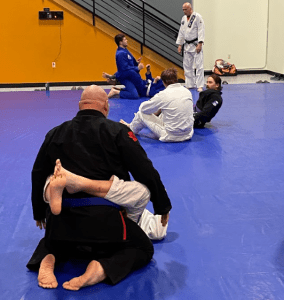 Jiu-Jitsu class with multiple participants practicing grappling techniques on a blue mat, under instructor supervision.