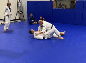 Jiu-Jitsu class with students practicing ground techniques on a blue mat, while other participants watch and smile.