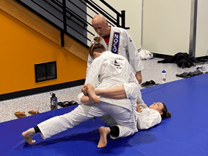 Brazilian Jiu Jitsu class with instructor observing two students practicing grappling techniques on a blue mat in the dojo.