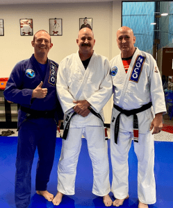 Three Brazilian Jiu-Jitsu practitioners in gis posing together on the mat, with two instructors and a student in the center.