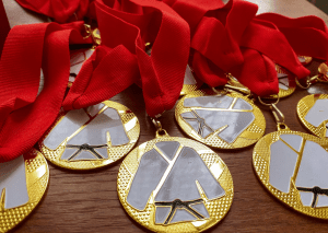 Close-up of gold medals with red ribbons, featuring a judo gi and black belt design, arranged on a wooden surface.