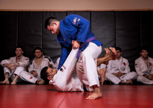 BJJ practitioner in a blue GI executing a guard pass while students observe closely during a training session.