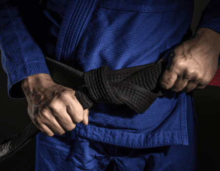 Close-up of a person in a blue gi tying a black belt, symbolizing dedication and progress in jiu-jitsu training.