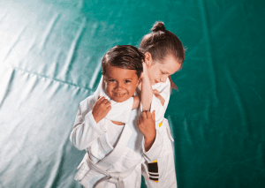 Two young children in white gis practicing jiu-jitsu on the mat, smiling and having fun during their training session.