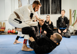 Two jiu-jitsu practitioners demonstrate techniques on the mat while others watch attentively in a training session.