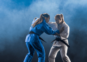 Two women in judo gi engaged in a grappling stance during training, showcasing focus and technique on a misty background.