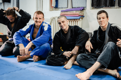 jiujitsuschools-1 A group of smiling practitioners in jiu-jitsu schools sit on mats wearing gis, showing camaraderie and joy.